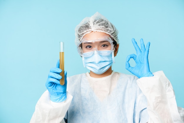 Laboratory worker nurse in personal protective equipment showing sample in test tube and okay sign t...