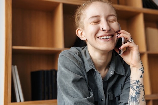 lachen vrij jong meisje met dreadlocks en piercing binnenshuis in de buurt van plank praten via de mobiele telefoon.