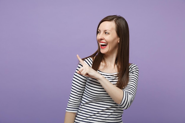 Lachen vrolijke jonge brunette vrouw in casual gestreepte kleding poseren geïsoleerd op violet paarse achtergrond studio portret. Mensen levensstijl concept. Bespotten kopie ruimte. Wijsvinger opzij wijzen.