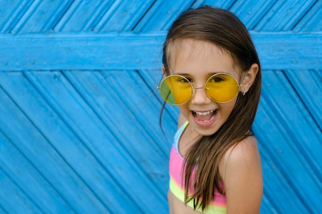 Lachend brunette meisje in gele zonnebril en een veelkleurig zwempak staat tegen een blauwe muur