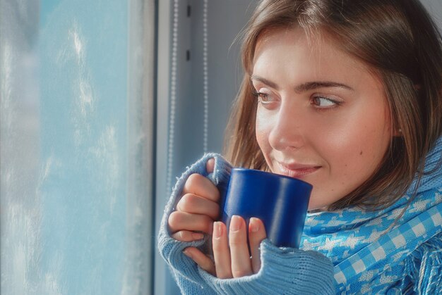 lachend dromend meisje in trui met de beker bij het bevroren raam in de winter