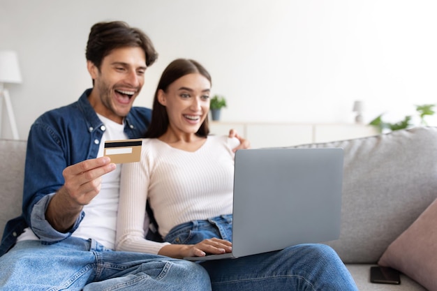 Lachend jong Europees koppel op de bank met creditcardlaptop en online winkelen in de woonkamer