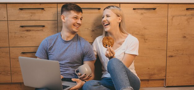 Lachend kaukasisch paar koekjes eten en samen met behulp van een laptop in de keuken op de vloer