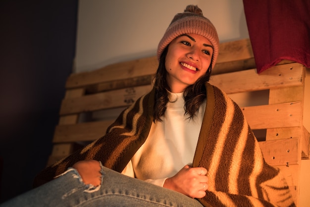 Lachend latina meisje met een winterhoed en een gescheurde spijkerbroek, gewikkeld in een winterdeken met een pallet erachter