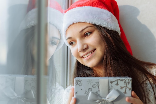 Foto lachend meisje in een kerstmuts met een geschenkdoos bij het raam close-up