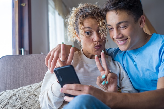 Lachende 40-jarige moeder rust met volwassen zoon die smartphone samen gebruikt, gelukkige jonge man geniet van familieweekend met moeder die een selfie neemt, veel plezier thuis