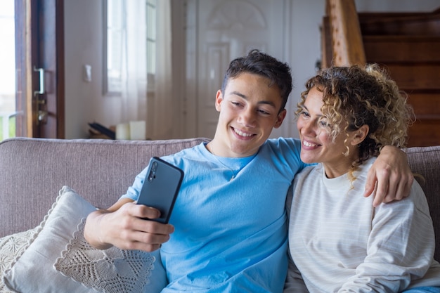 Lachende 40-jarige moeder rust met volwassen zoon die smartphone samen gebruikt, gelukkige jonge man geniet van familieweekend met moeder die een selfie neemt, veel plezier thuis