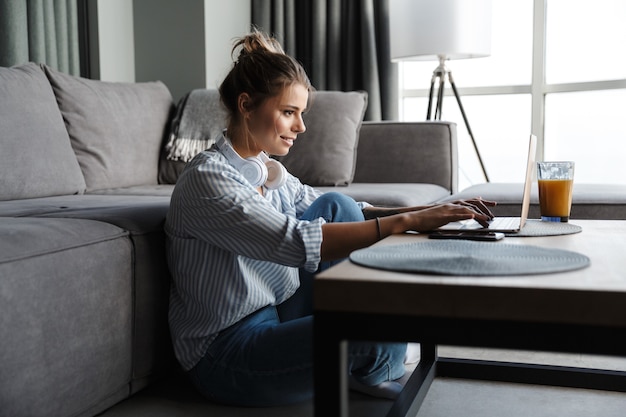 Foto lachende aardige vrouw met koptelefoon die laptop gebruikt terwijl ze op de vloer in de woonkamer zit