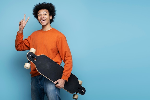 Lachende Afro-Amerikaanse man met longboard met overwinningsteken geïsoleerd op blauwe achtergrond