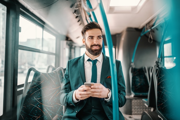 Lachende ambitieuze bebaarde zakenman in turquoise pak zitten in de openbare bus en het gebruik van slimme telefoon. Als je wilt vliegen, geef dan alles op dat je zwaar maakt.