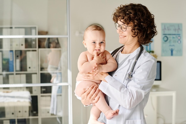Foto lachende arts in witte jas die baby op de armen houdt terwijl hij op kantoor staat