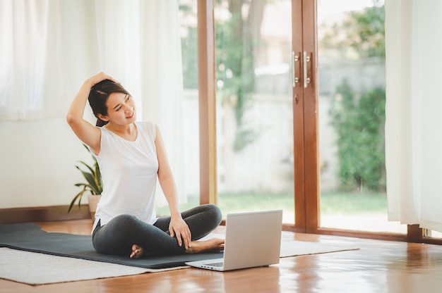 Lachende Aziatische vrouw doet yoga nek uitrekken online klas van laptop thuis in de woonkamer.