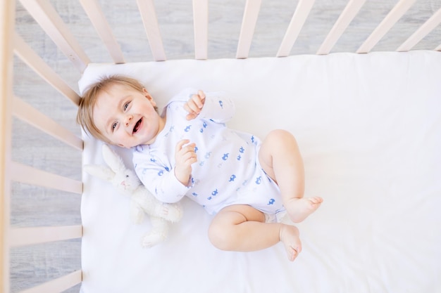 Lachende baby blonde jongen ligt in een wieg op zijn rug in een kamer thuis met blauw en wit katoenen beddengoed en glimlacht in de ochtend het concept van kinderartikelen en accessoires