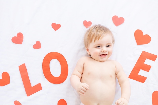 Lachende baby blonde jongen ligt op een bed met rode harten en de inscriptie liefde thuis en glimlacht in de ochtend het concept van Valentijnsdag of Moederdag