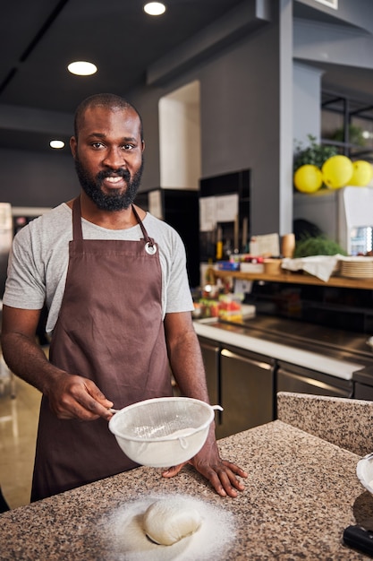 lachende bebaarde man in schort met zeef voor deeg tijdens het koken van bakkerij in de keuken