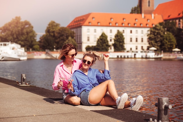 Lachende beste vrienden meisjes drinken wijn op een wandeling portret van twee gelukkige meisjes romantisch zittend op rivwr achtergrond met fles in de hand praten en glimlachen emoties concept