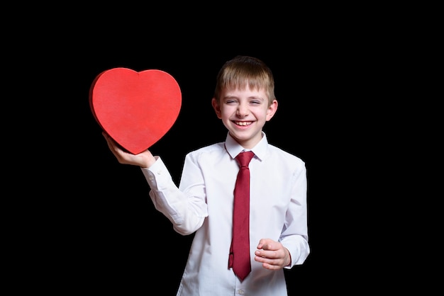 Lachende blonde jongen in een overhemd en stropdas met een rode hartvormige doos. Liefde en familie concept.