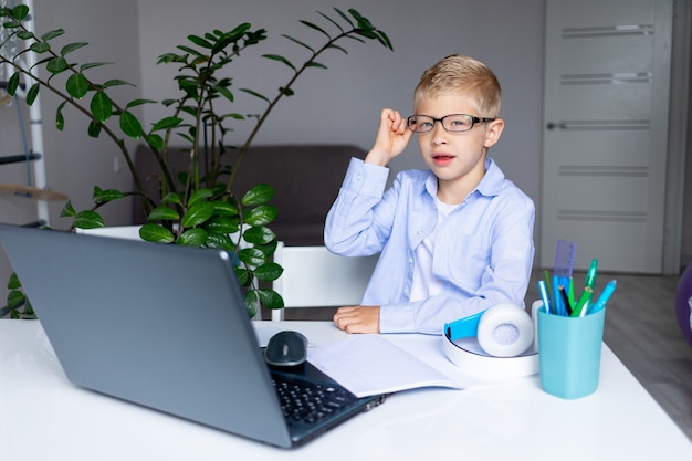 Lachende blonde jongen met een bril maakt een videogesprek via laptop tijdens afstandsonderwijs thuis