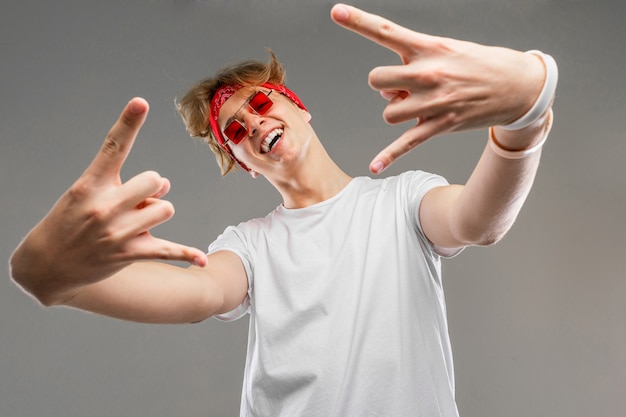 lachende blonde man met een bandana in glazen en een wit t-shirt toont een rocker-teken met zijn handen naar voren op een grijze muur