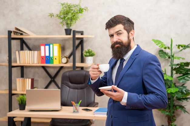 Lachende brutale ceo in zakelijk pak heeft lunchpauze met koffiekopje bij de kantoorondernemer