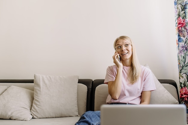 Foto lachende casual freelancer met behulp van slimme telefoon en laptop zittend op de bank.