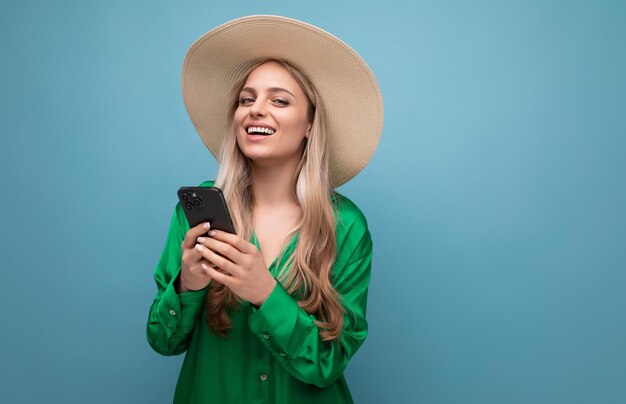 Lachende charmante jonge volwassen vrouw in een zomerhoed met een telefoon in haar handen op een blauwe achtergrond