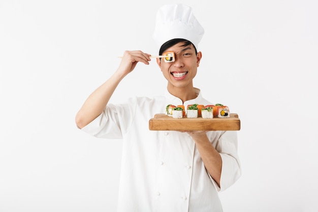 lachende chinese chef-man in het wit kok uniform bedrijf plaat en het eten van sushi set met stokjes geïsoleerd over witte muur