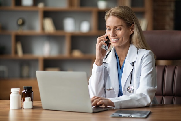 Lachende dokter vrouw praten op mobiele telefoon en met behulp van Laptop op de werkplek