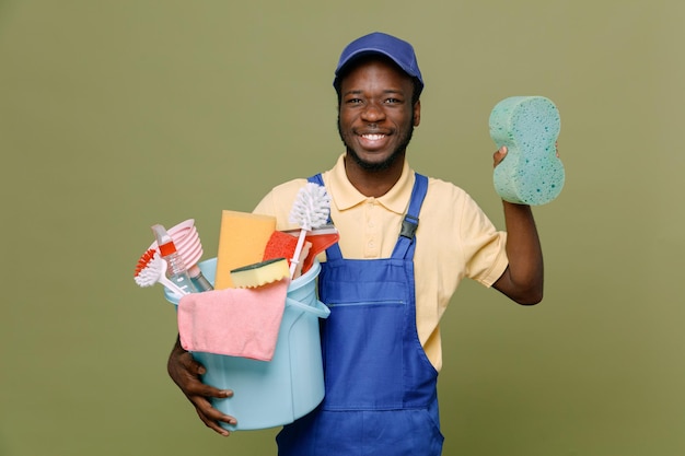 Lachende emmer met schoonmaakmiddelen met spons jonge Afro-Amerikaanse schonere man in uniform met handschoenen geïsoleerd op groene achtergrond