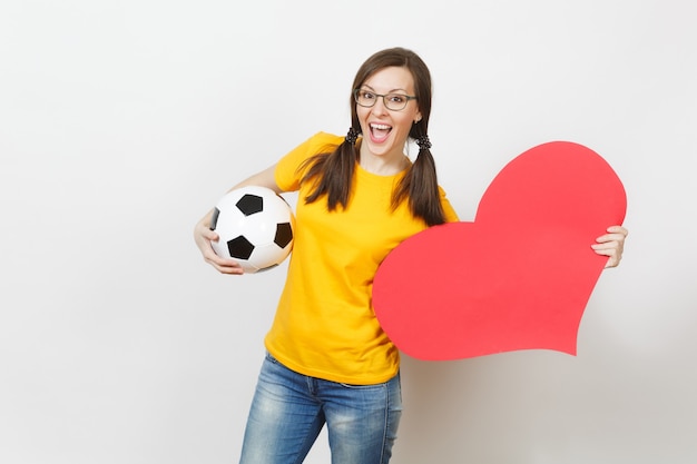 Lachende Europese vrouw, leuke paardenstaarten, voetbalfan of speler in glazen, gele uniform houden klassieke voetbal, rood hart geïsoleerd op een witte achtergrond. Sport, voetbal, gezond levensstijlconcept.