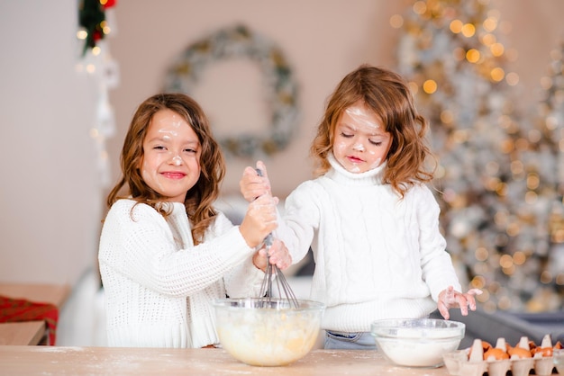 Lachende gelukkige jongen meisjes koken zelfgemaakt cakedeeg in glazen kom met meel en eieren boven kerstboomverlichting en decoraties op keukentafel thuis Winter vakantieseizoen Jeugd