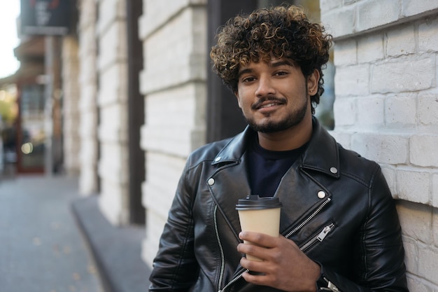 Foto lachende indiase man met zwarte leren jas die een kopje koffie vasthoudt terwijl hij naar de camera kijkt