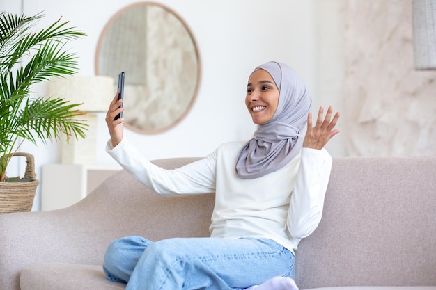 Lachende jonge arabische vrouw in hijab die thuis in het hotel op de bank zit en aan de telefoon praat