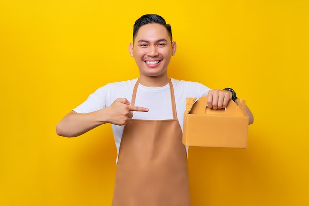 Lachende jonge Aziatische man barista barman werknemer dragen bruine schort werk in coffeeshop wijzende vinger op blanco ambachtelijk papier afhaalmaaltijden cake tas op gele achtergrond Opstarten van kleine bedrijven