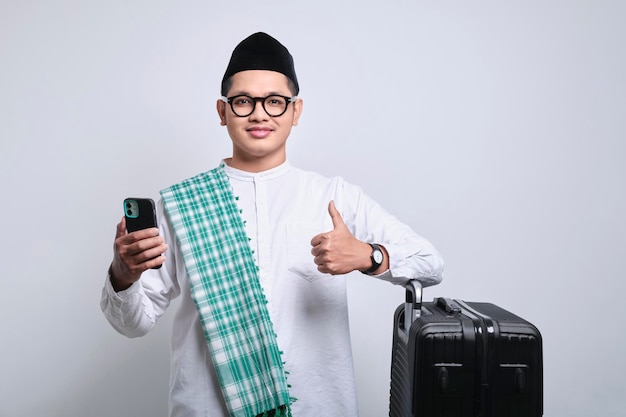 Lachende jonge Aziatische moslim man duim omhoog gebaar tonen terwijl smartphone en leunend op koffer klaar voor de vakantie Ramadan en eid Mubarak concept