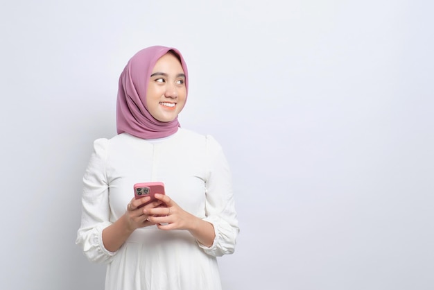 Lachende jonge Aziatische moslimvrouw die mobiele telefoon gebruikt en wegkijkt naar kopie ruimte geïsoleerd op witte achtergrond