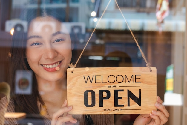 Lachende jonge aziatische ondernemer, medewerker detailhandel, coffeeshopvrouw, draaiend meisje, uithangbord instellen om te openen voor welkome klant, winkel heropenen na sluiting quarantaine in covid. labelconcept.