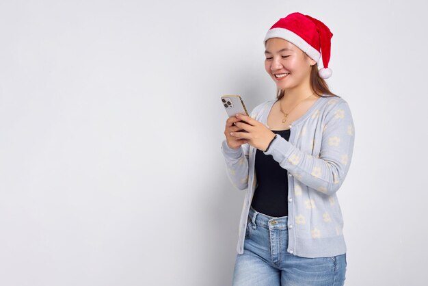 Lachende jonge Aziatische vrouw in een kerstmuts kijkend naar smartphone die geweldig nieuws krijgt geïsoleerd op witte achtergrond