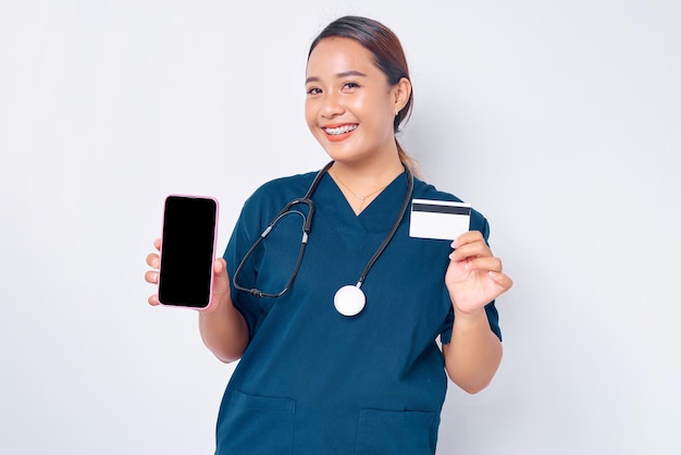 Lachende jonge Aziatische vrouw professionele verpleegster die werkt in een blauw uniform met een leeg scherm mobiele telefoon en een creditcard op een witte achtergrond houdt Gezondheidszorg geneeskunde concept