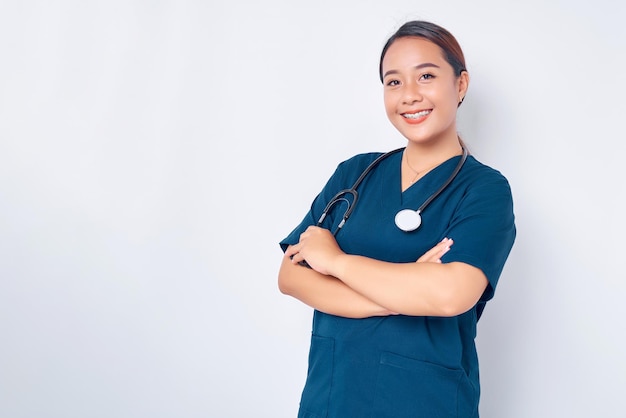 Lachende jonge Aziatische vrouw verpleegster dragen blauw uniform met stethoscoop kruis armen borst en kijken naar de camera geïsoleerd op witte achtergrond Gezondheidszorg geneeskunde concept