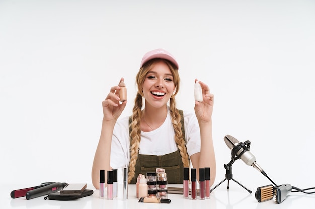 Lachende jonge blonde meid blogger die een schoonheidsvideo maakt met make-up terwijl ze aan de tafel zit geïsoleerd op wit