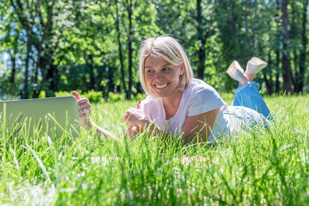 lachende jonge blonde vrouw ligt op het gras in een park met een laptop op een zonnige zomerdag.