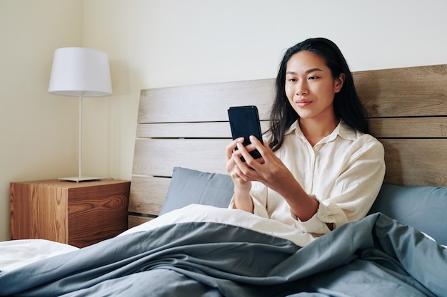 Lachende jonge Chinese vrouw zittend in bed na het ontwaken en het lezen van SMS-berichten op haar smartphone