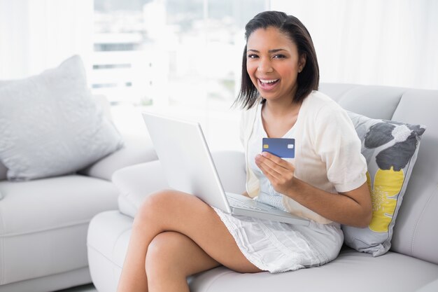 Lachende jonge donkerharige vrouw in witte kleren online winkelen met een laptop