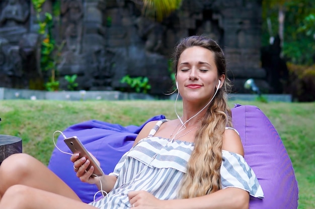 Lachende jonge langharige vrouw rust op terras in het park en luistert naar muziek op de smartphone het concept van moderne digitale levensstijl