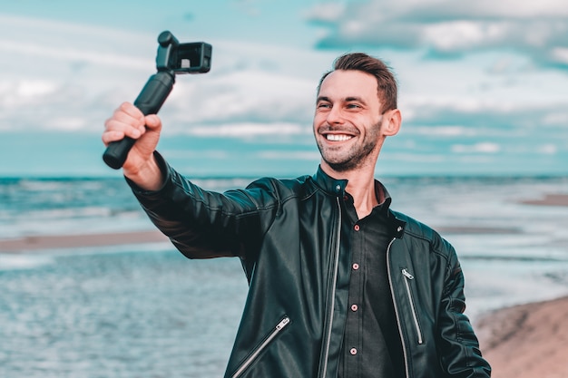 Lachende jonge mannelijke blogger selfie of streaming video maken op het strand met behulp van actiecamera met gimbal camera stabilizer.