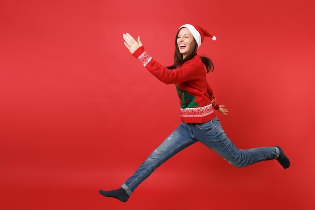 Lachende jonge Santa meisje in gebreide trui, kerstmuts springen, stijgende handen, benen spreiden geïsoleerd op rode achtergrond. Gelukkig Nieuwjaar 2019 viering vakantie partij concept. Bespotten kopie ruimte.