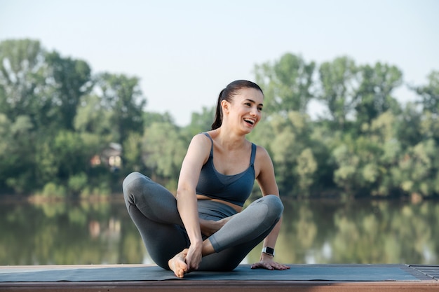 Lachende jonge vrouw die buiten yoga doet op een prachtige plek aan een rivier