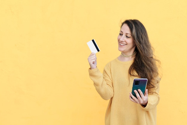 Lachende jonge vrouw die mobiele telefoon gebruikt met een bankcreditcard in de hand