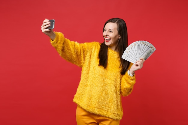 Lachende jonge vrouw die selfie schot op mobiele telefoon doet, fan van geld in dollarbankbiljetten houdt, contant geld geïsoleerd op rode achtergrond. mensen oprechte emoties, lifestyle concept. bespotten kopie ruimte.